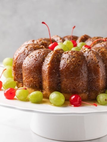 A rum bundt cake on a white cake stand garnished with green grapes and maraschino cherries