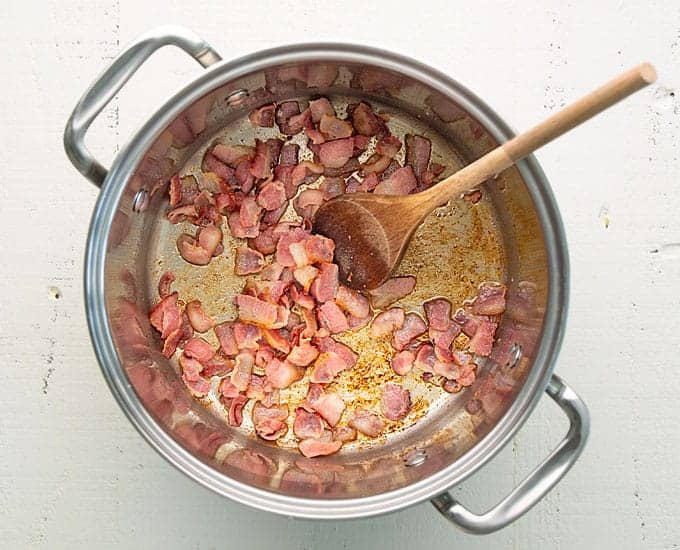 Overhead view of fried bacon pieces in a large stainless pot with a wooden spoon.