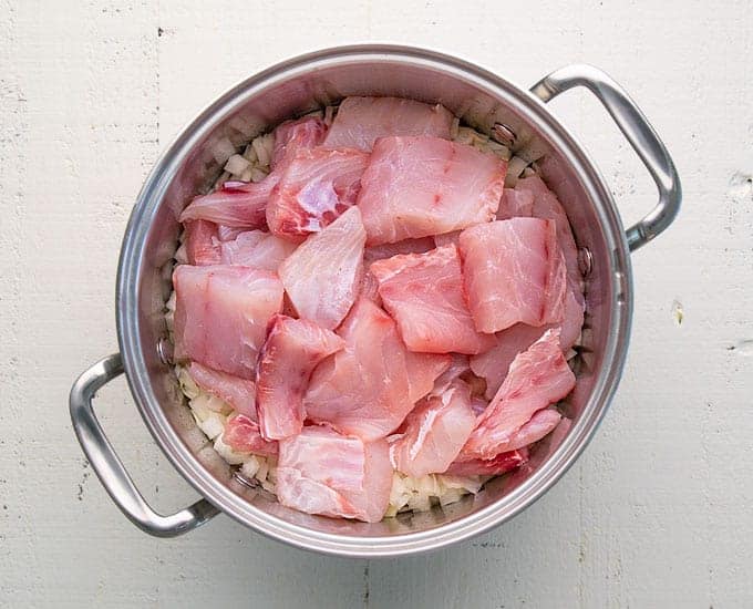 Overhead view of chunks of raw fish over onions and potatoes in a stainless pot.