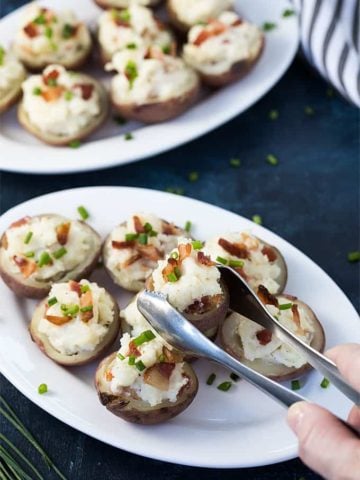 A pair of tongs holding a potato popper sprinkled with bacon and chives.