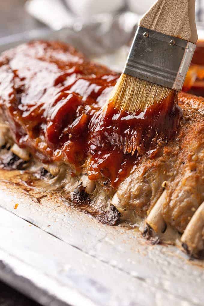 A rack of baked pork ribs on a baking sheet being brushed with barbecue sauce.