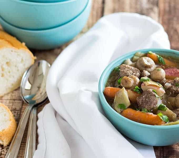 Beef stew in a blue bowl beside a white napkin, spoons and French bread.