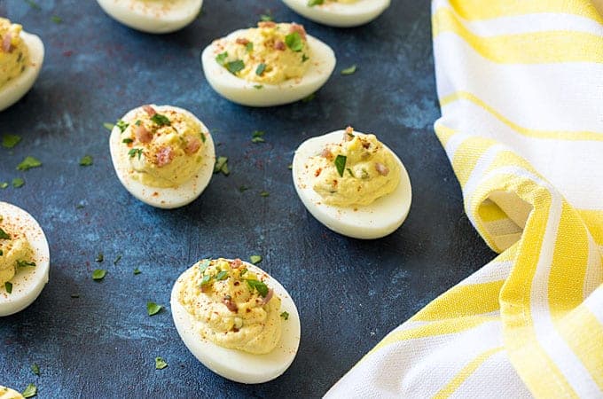 Overhead view of deviled eggs with horseradish and bacon on a blue surface by a striped napkin.