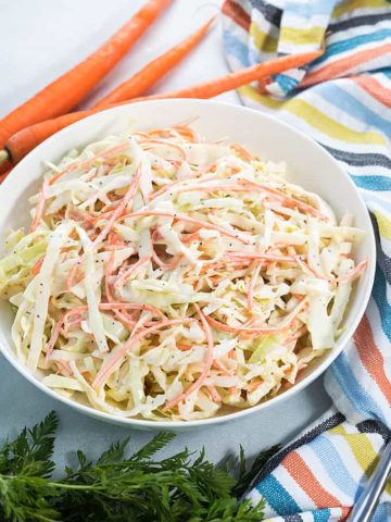 A white bowl of creamy Carolina coleslaw beside a striped napkin and three carrots.