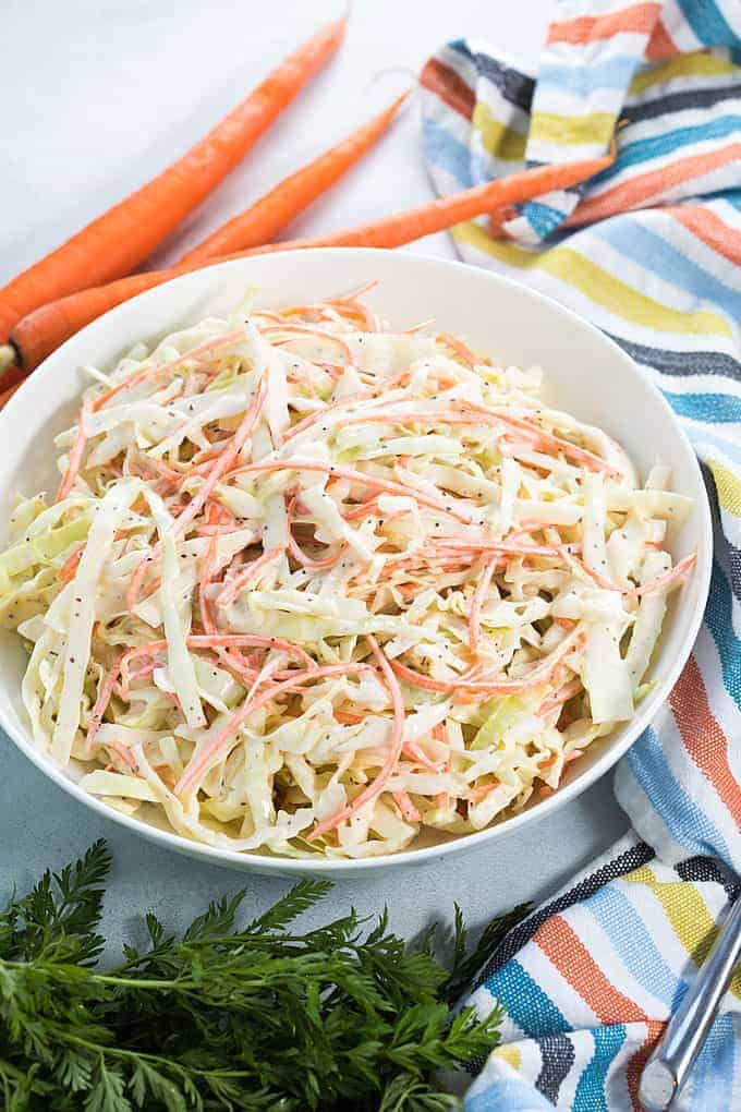 A white bowl of creamy Carolina coleslaw beside a striped napkin and three carrots.