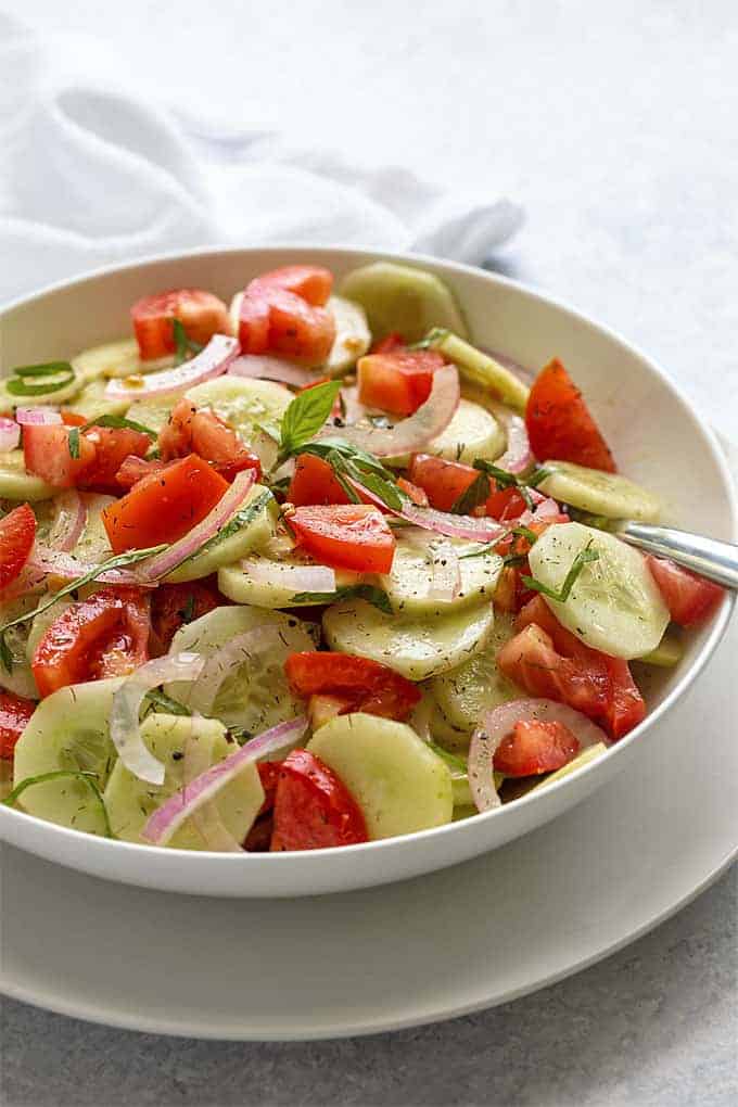 Cucumber and tomato salad in a white bowl with a serving spoon.