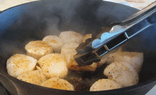 Scallops being seared in a cast iron skillet with tongs turning a scallop.