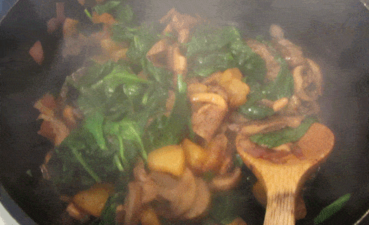 Closeup view of mushrooms, chopped pineapple and spinach in a skillet with a wooden spoon.