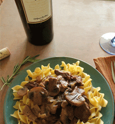 Overhead view of beef and mushrooms over noodles on a teal plate with a bottle of wine.