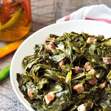 A white bowl of collard greens with pork pieces by a jar of vinegar.