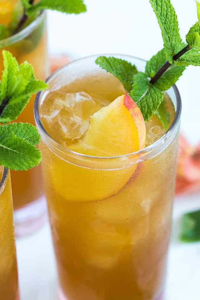 A closeup of a glass of iced peach tea with whiskey garnished with peaches and mint.