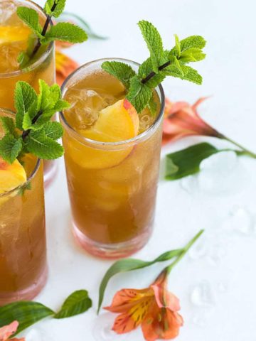 Three glasses of peach tea whiskey cocktail garnished with sliced peaches and mint.