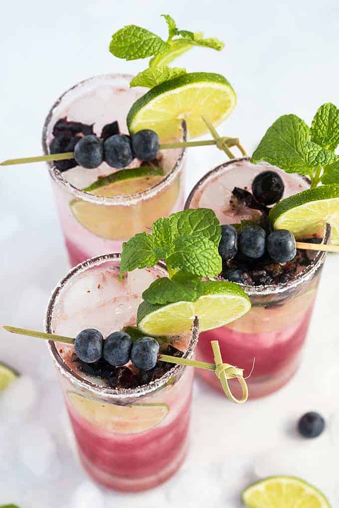 Overhead view of three mojitos garnished with blueberries, lime and mint in sugar-rimmed glasses.