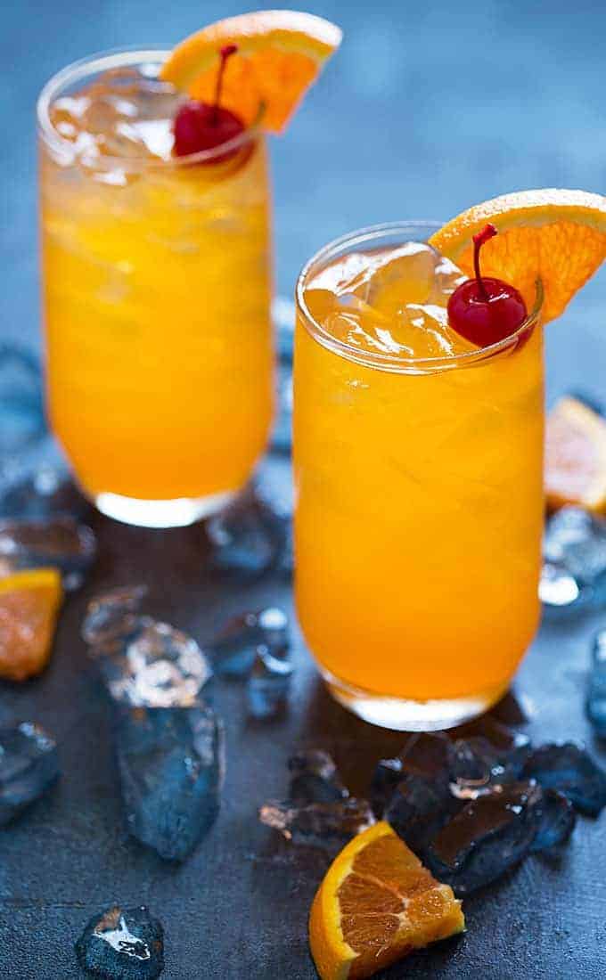 Two creamsicle vodka drinks garnished with cherries and orange slices on a blue surface.