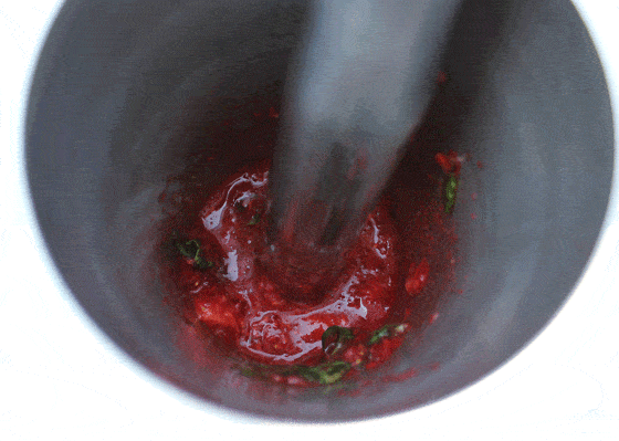 Overhead closeup of muddling strawberries and basil in a stainless cocktail shaker.