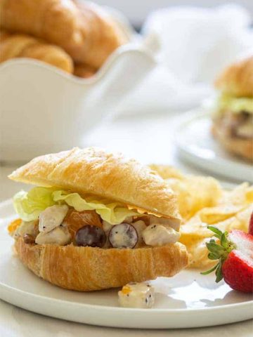 Summer chicken salad on a croissant with fresh strawberries and potato chips on a round white plate