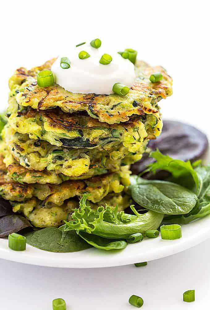 A stack of zucchini fritters topped with sour cream and green onions on a white plate.
