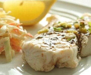 A closeup of baked fish, coleslaw and lemon wedge on a white plate.