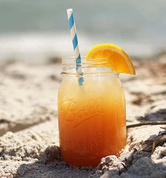 An orange cocktail in a mason jar garnished with orange on sand on the beach.