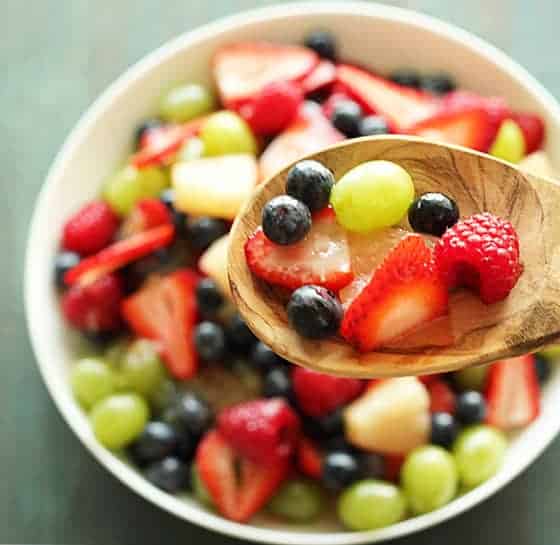 Overhead view of a wooden spoon holding fruit salad over a white bowl.