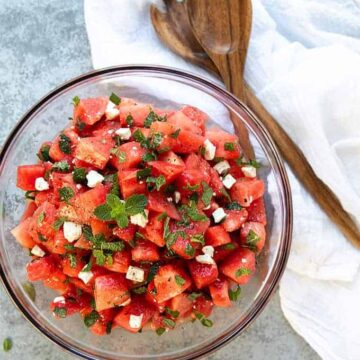 Overhead view of watermelon feta salad with mint in a glass bowl beside wooden salad servers