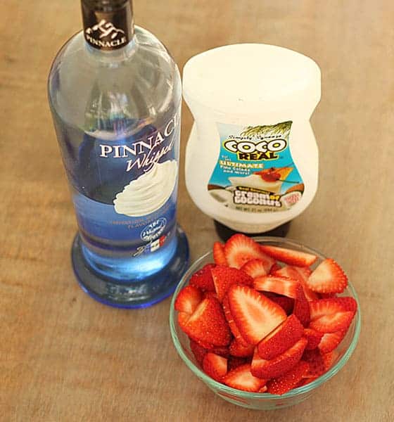 A bottle of vodka, container of cream of coconut and small bowl of sliced strawberries.