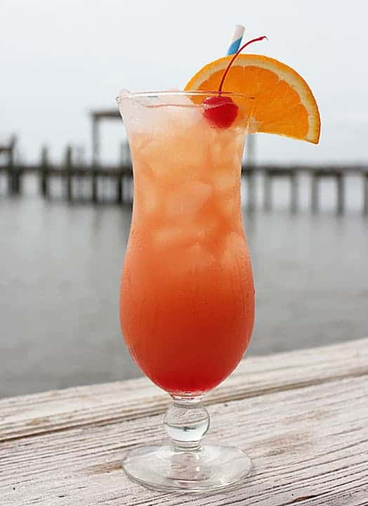 An orange cocktail in a hurricane glass garnished with orange and a cherry.  Water is in the background.