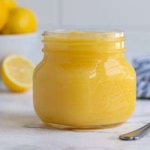 A jar of lemon curd with a bowl of lemons in the background.
