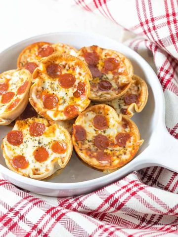 Mini appetizer pepperoni pizzas in a white dish beside a checked towel.