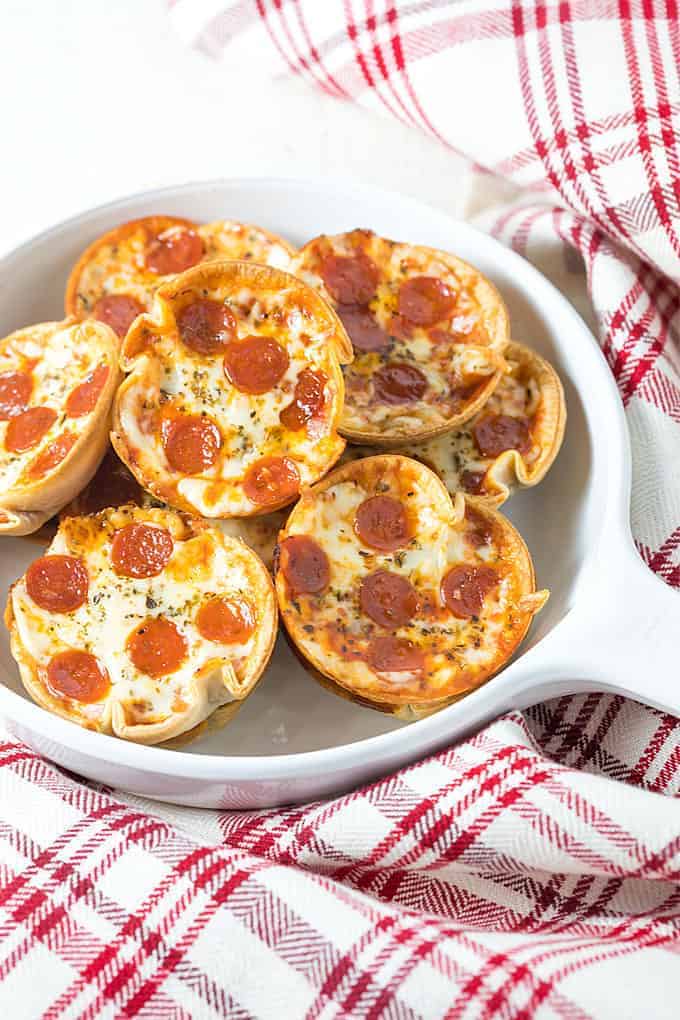 Mini appetizer pepperoni pizzas in a white dish beside a checked towel.