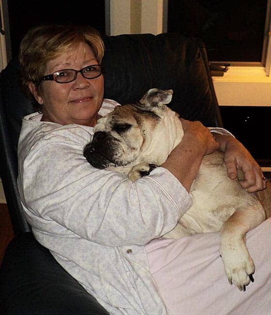 A woman with brown short hair in a chair wearing lounge clothes hugging a bulldog.