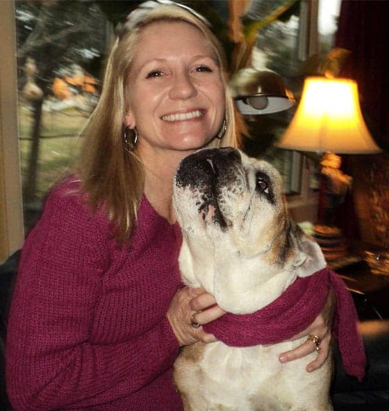 A young woman with blonde hair smiling with a bulldog in her lap.