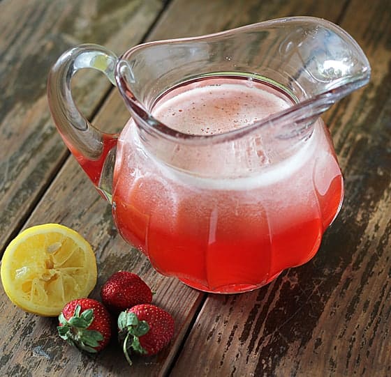 Strawberry lemonade in a glass pitcher on a wooden surface.