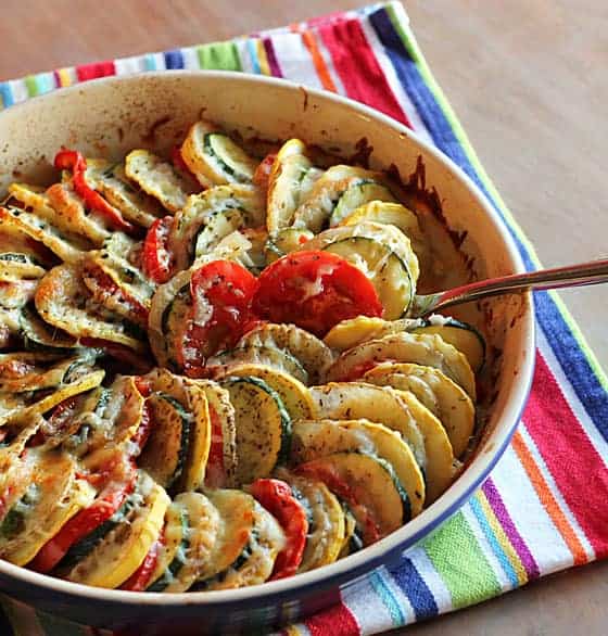 Baked thinly sliced squash, tomatoes and potatoes topped with cheese in a baking dish with a spoon.