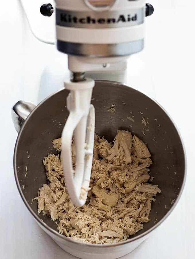 Shredded chicken in a mixing bowl of a white stand mixer with the paddle attachment.