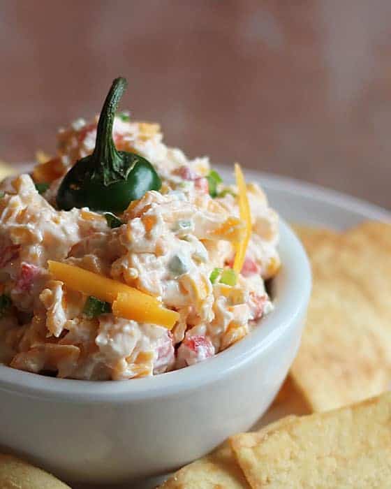 Pimento cheese topped with a jalapeno stem in a white bowl surrounded by chips.