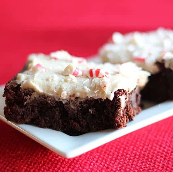 Brownies topped with icing and crushed peppermint on a white plate on a red background.