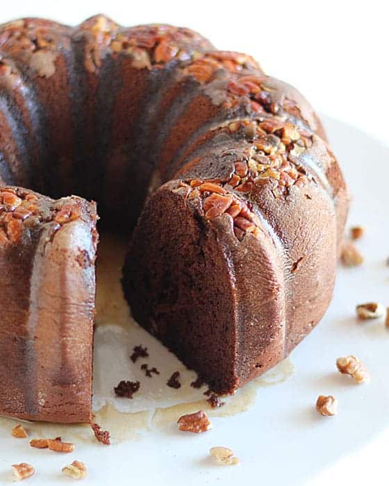 A chocolate bundt cake topped with pecans and glaze on a white plate.