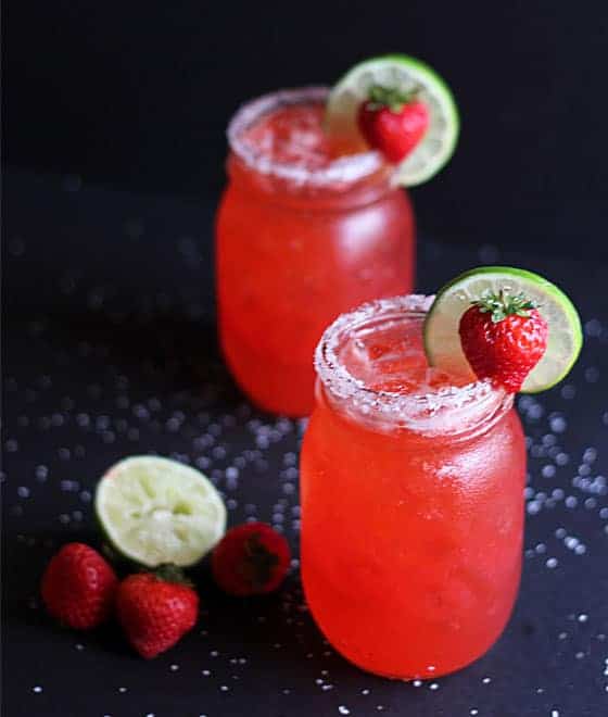Two garnished strawberry margaritas in mason jars on a black background.