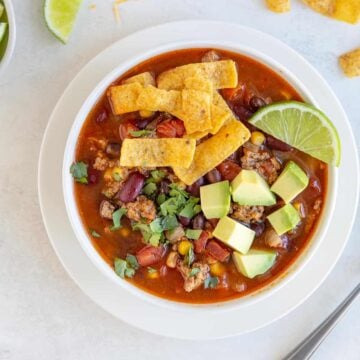 A white bowl of taco soup with corn chips, avocado and a lime wedge.