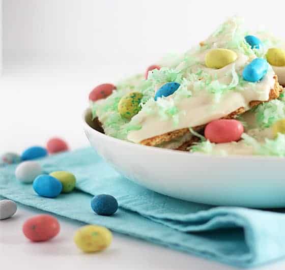 Front closeup view of graham cracker pieces topped with white chocolate, robin candy eggs and green coconut in a white bowl.