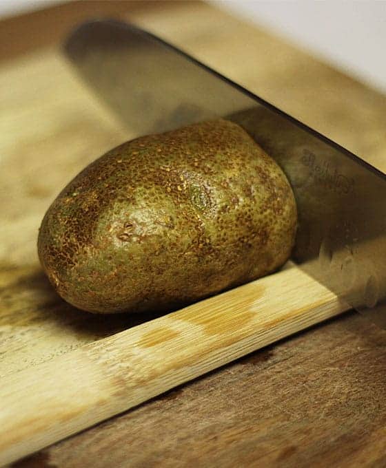 A knife cutting through a baking potato with a wooden spoon handle in front of the potato.