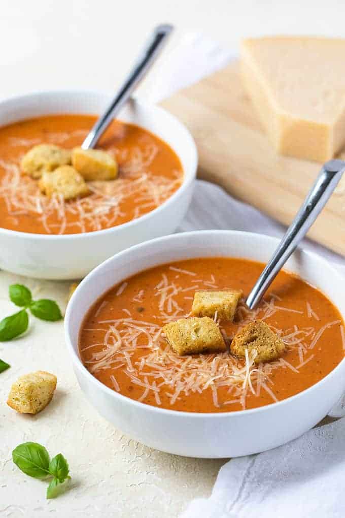 Two white bowls of soup topped with Parmesan and croutons. Spoons are in each bowl.