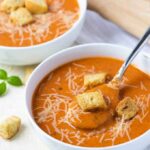 A spoon in a white bowl of tomato soup topped with grated Parmesan cheese and croutons.