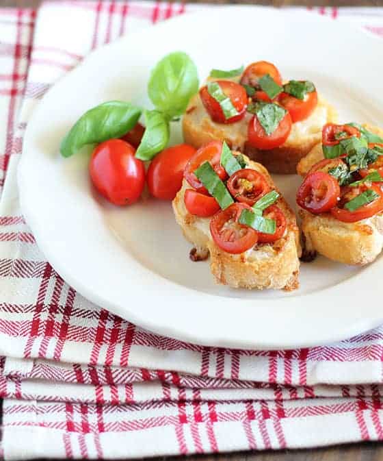 A closeup view of 3 baguette slices topped with cheese, sliced cherry tomatoes and basil on a plate.