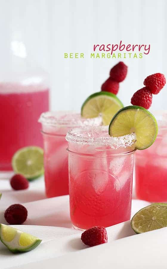 Dark pink colored margaritas in small jars garnished with fresh raspberries and lime on a skewer.