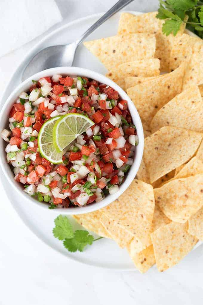 Overhead view of Pico de Gallo in a white bowl on a plate with tortilla chips.