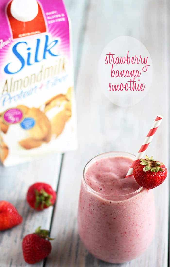 A smoothie with a striped paper straw and strawberry garnish.  A carton of almondmilk is in background.
