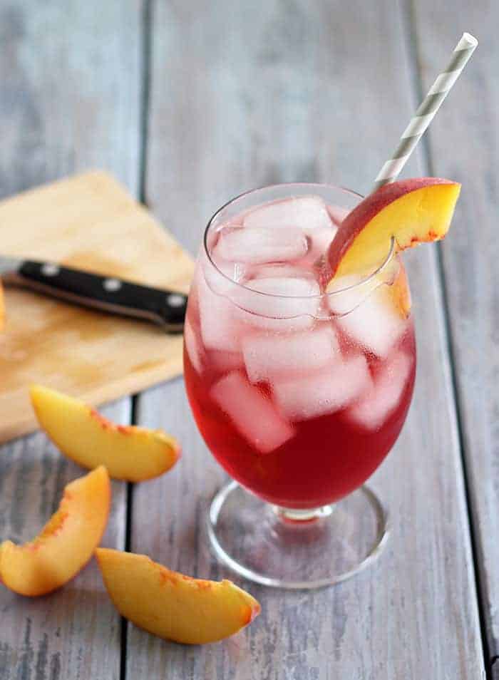 A cocktail prepared with peach vodka, cranberry juice and club soda beside fresh sliced peaches.