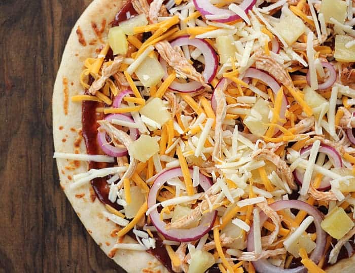 Overhead view of an unbaked pizza with barbecue sauce, cooked chicken, pineapple, cheese and sliced onion.